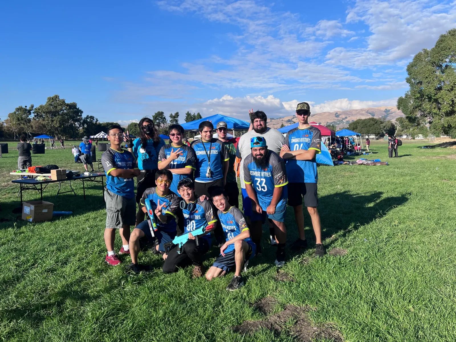 Twelve teammates in blue and grey Combat Kitties jerseys posed together on a grass field at an outdoor tournament, two of them holding blue blasters