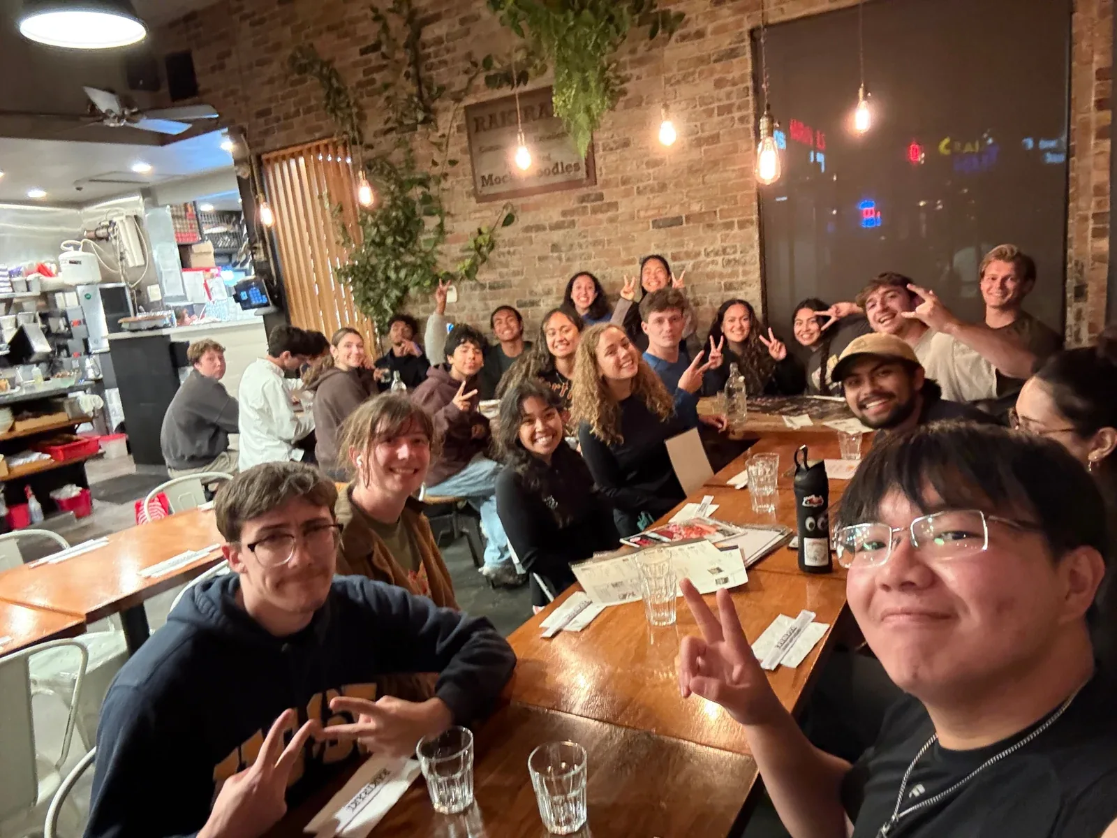 A group seated along long tables at a team meal, the nearest few waving at the camera